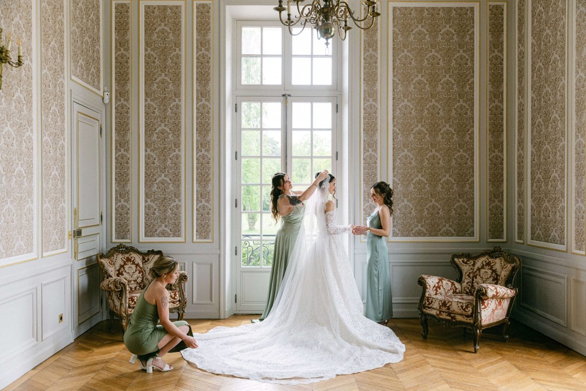 Bride in a lace wedding gown with a long train as two bridesmaids adjust her veil in an elegant, wallpapered room.