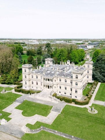 A large stone castle-like mansion with symmetrical towers and manicured gardens, viewed from above.