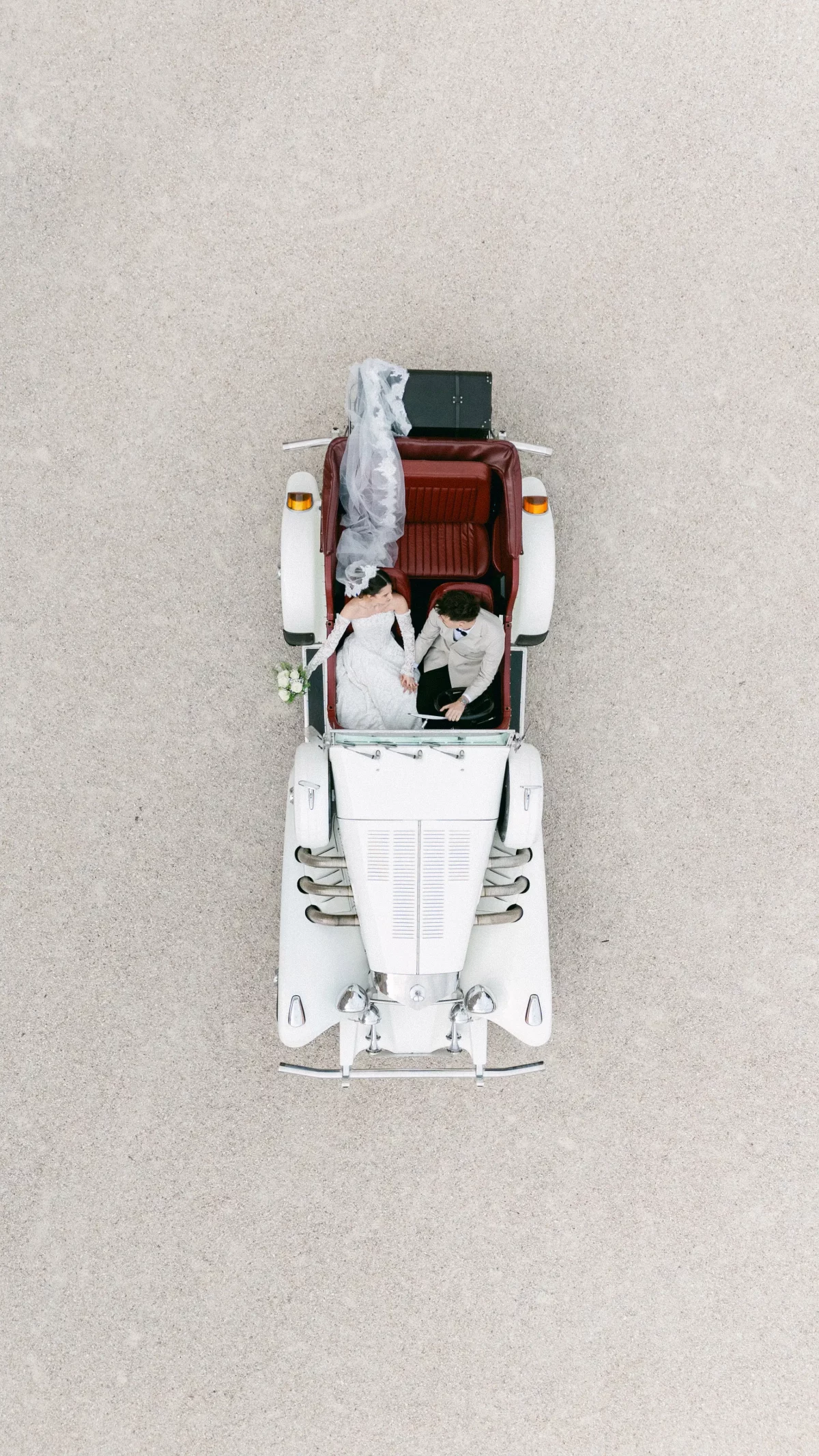 Top-down view of a white vintage convertible with red seats; bride and groom sit in the back, bride's veil flowing and bouquet visible on the side.
