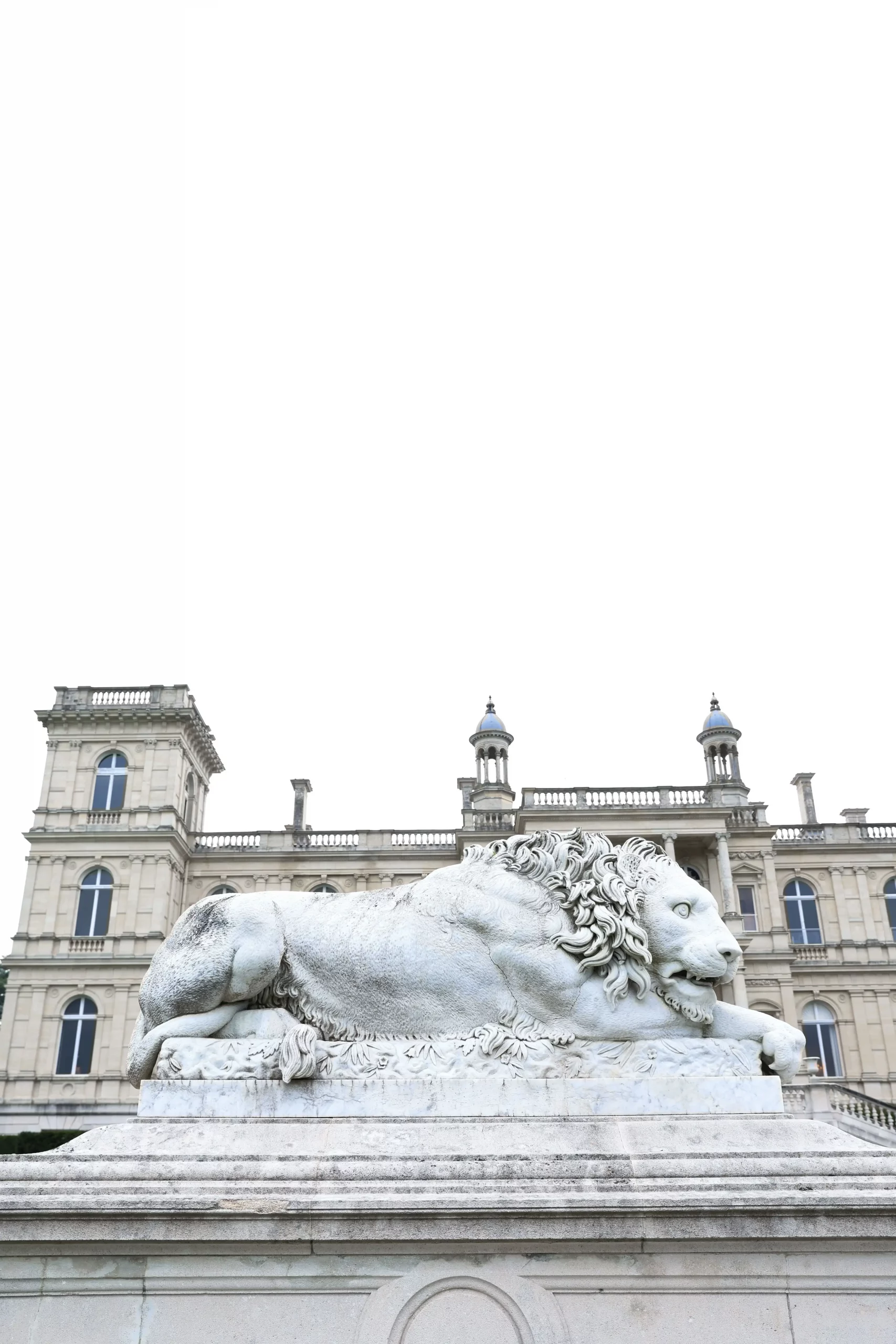 Lying stone lion statue on a pedestal in front of a grand historic building with arched windows and decorative towers.