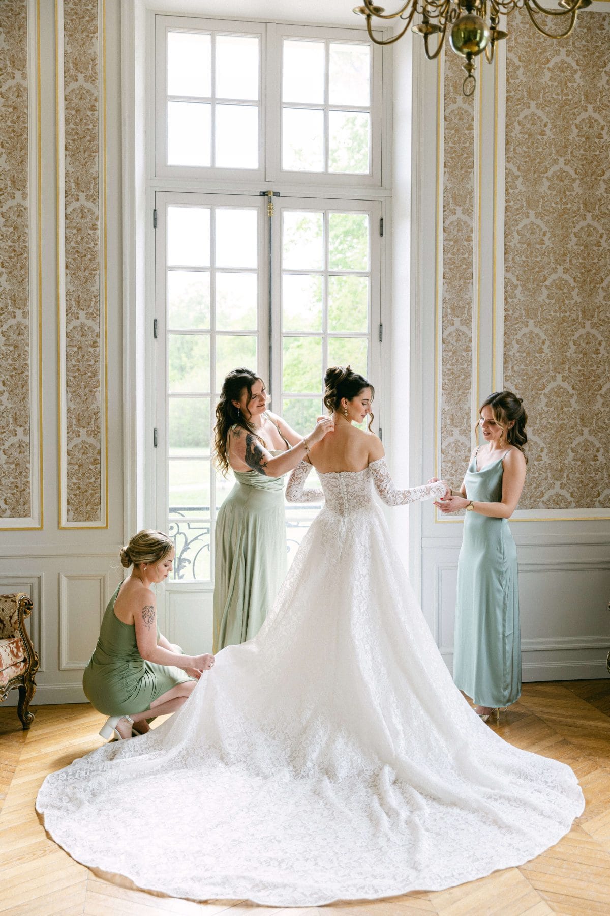 Mariée-chateau-de-nainville-demoiselles-honneur | Séverine Alves - Paris wedding photographer, France Bride in a white lace wedding gown with a long train being helped by three bridesmaids in pale sage dresses.