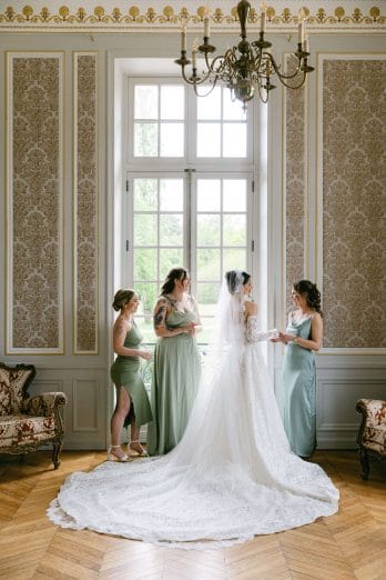 Photographe-mariage-Paris11 | Séverine Alves - Paris wedding photographer, France Bride in a white lace wedding gown and veil stands with two bridesmaids in sage green dresses in an ornate room by a large window.