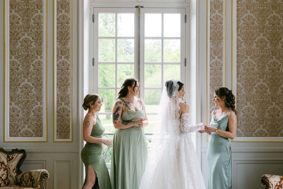 Bride in a white lace wedding gown and veil stands with two bridesmaids in sage green dresses in an ornate room by a large window.