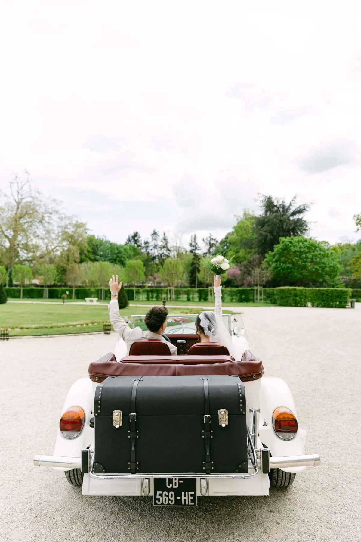 Bride and groom in a vintage white convertible drive away, arms raised in celebration; bouquet visible in the bride's hand/seat.