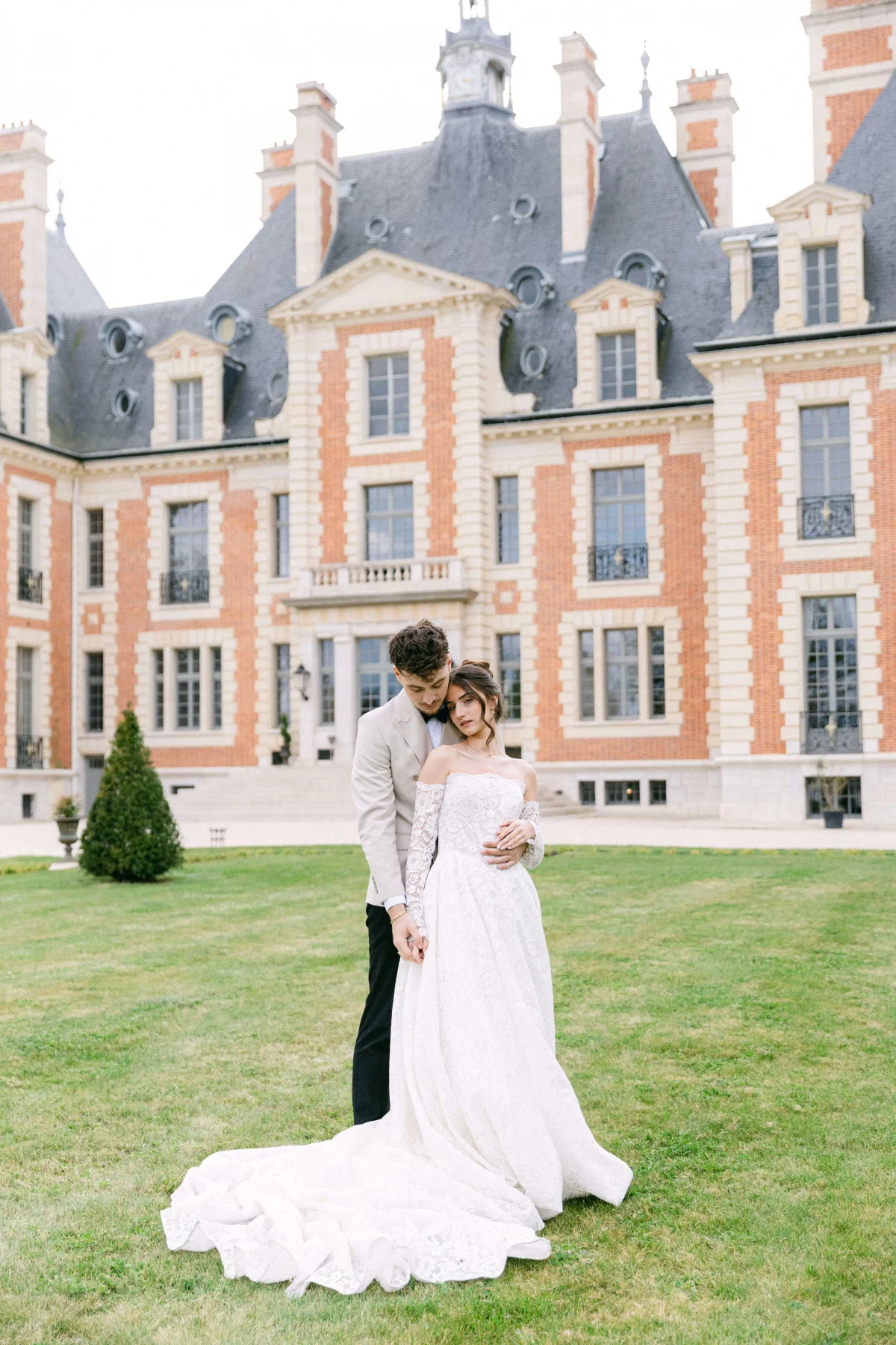 Bride and groom embrace on a green lawn in front of a red-brick château with slate roof, the bride in an off-shoulder lace gown and long train.