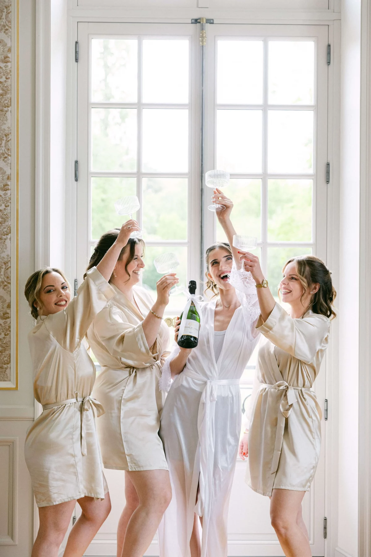 Bride in a white robe toasting with four bridesmaids in beige robes by a large window, holding champagne glasses and a bottle of bubbly.