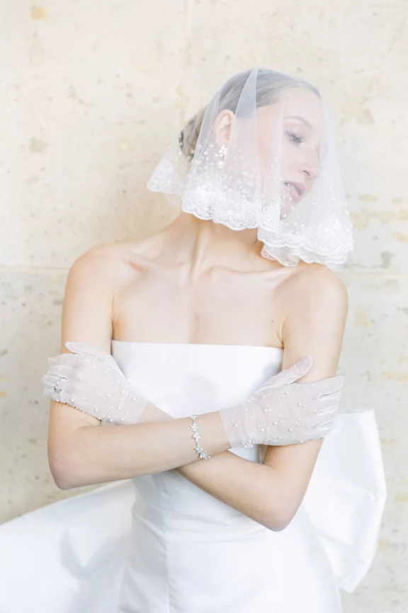 Bride in a white strapless gown with pearl-embellished gloves and a lace-edged veil, arms crossed.