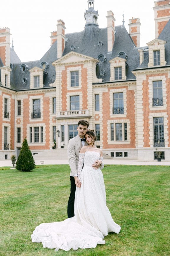 Bride in a lace wedding gown and groom in a beige suit embrace on a green lawn in front of a grand red-brick manor with tall chimneys for a wedding photo.