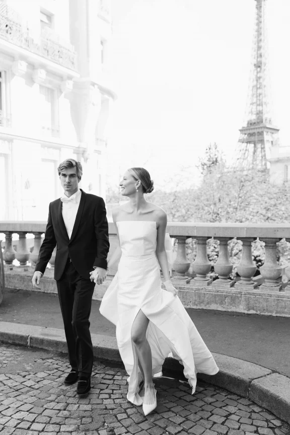 Bride in a strapless white gown and a groom in a black suit walk hand in hand along a Paris street, with the Eiffel Tower in the background.