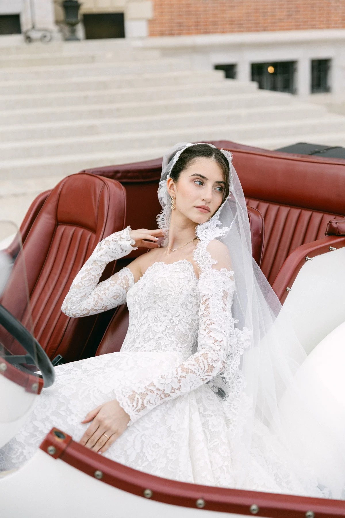 Bride in a lace wedding dress and veil seated in a red vintage car, gazing to the right.