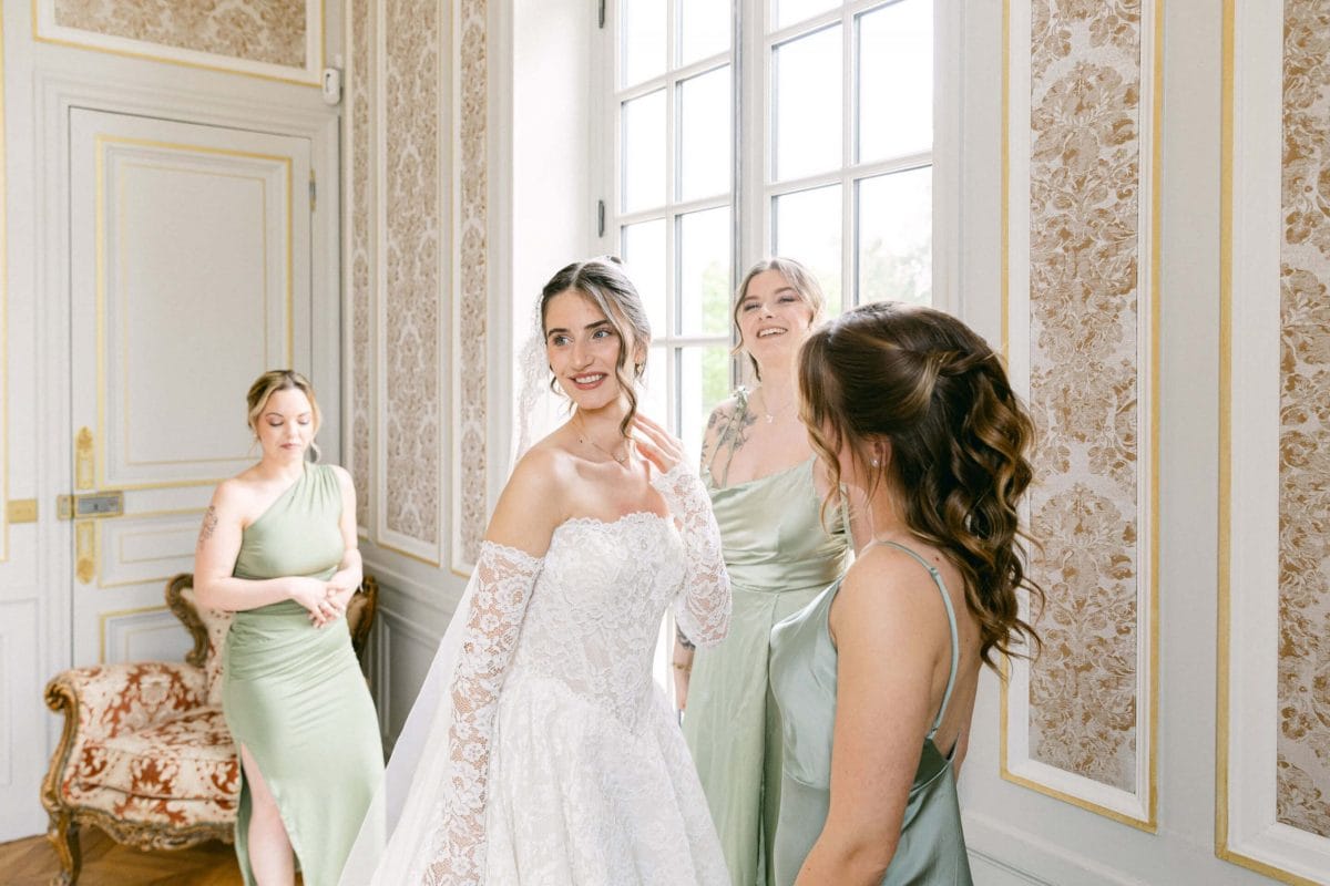Bride in a lace wedding dress smiles as bridesmaids in sage gowns help her in an ornate sunlit room with gold-trimmed wallpaper and tall windows to the side.